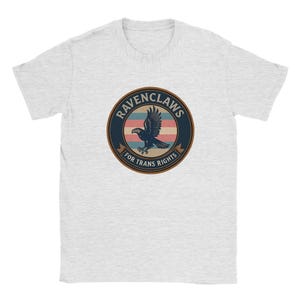 Pode incluir: Uma t-shirt cinzenta clara de manga curta com um gráfico circular no peito. O design inclui uma águia azul-escura em voo sobre riscas horizontais azul-claras, brancas e cor-de-rosa, com o texto RAVENCLAWS FOR TRANS RIGHTS.