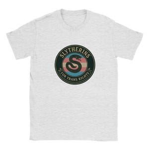 May include: A light gray short-sleeve t-shirt featuring a circular graphic on the chest. The design includes a black snake coiled into an S shape, centered over horizontal pink, white, and blue stripes, with the text SLYTHERINS FOR TRANS RIGHTS.