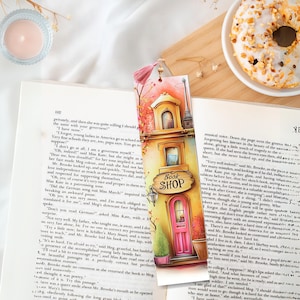 May include: A colorful bookmark featuring a whimsical illustration of a bookshop with a pink door and the text "Book Shop." The bookmark is set against a backdrop of an open book and a glazed donut.