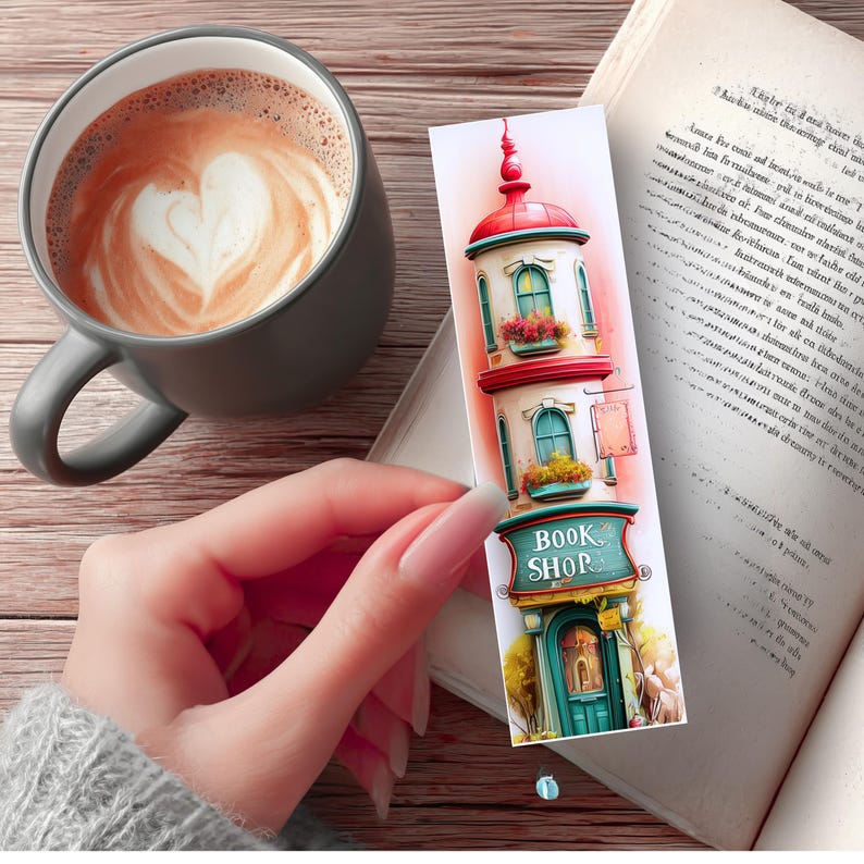 May include: A colorful bookmark featuring a whimsical illustration of a book shop with the text "BOOK SHOP". The bookmark is held near an open book and a gray mug of coffee with a heart-shaped design in the foam.