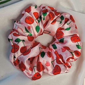 Puede incluir: Una scrunchie de satén rosa con un estampado repetido de cerezas rojas sonrientes, hojas verdes y pequeños corazones rosas. Este accesorio para el cabello es ideal para añadir un toque divertido a cualquier peinado.