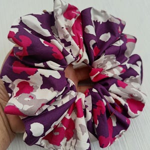Peut inclure: Un grand chouchou avec un motif floral vibrant dans les tons de violet, rose, blanc et gris. Le tissu est froncé et plissé, créant un accessoire volumineux et texturé. Cet accessoire pour cheveux est conçu pour maintenir les cheveux en queue de cheval ou en chignon.