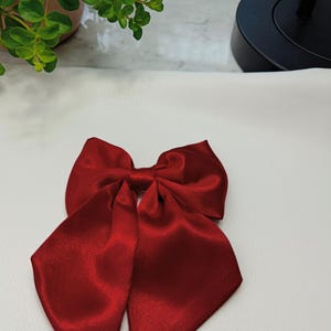 Puede incluir: Un lazo grande de satén rojo con colas largas y puntiagudas. El lazo está hecho de una tela brillante y lisa y está centrado en una superficie blanca. El lazo es de un color rojo intenso y rico.