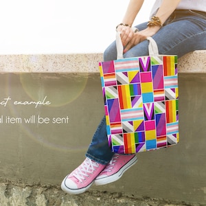 Puede incluir: Un bolso tote colorido con un dise&ntilde;o geom&eacute;trico que incluye los colores de la bandera del orgullo transg&eacute;nero. El bolso, con asas blancas, lo sostiene una persona que lleva vaqueros y zapatillas rosas. El texto de la imagen dice: "product example, no physical item will be sent."