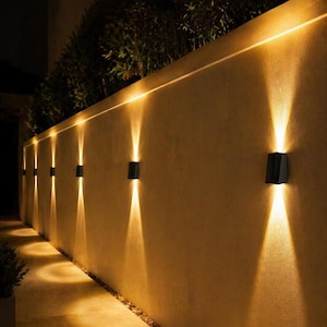 Puede incluir: Luces de pared exteriores negras y rectangulares iluminan una pared beige con un brillo cálido. Las luces emiten un haz de luz doble. Una luz lineal recorre la parte superior de la pared. Se ve follaje verde.