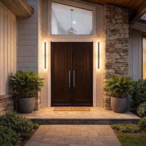 Pode incluir: Vista exterior da entrada de uma casa com uma porta dupla castanha escura. A entrada é ladeada por colunas de pedra e luzes verticais. Plantas em vasos e um caminho de tijolos conduzem à porta.