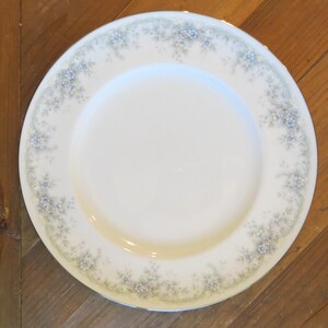 May include: White ceramic plate with a floral border featuring blue and yellow flowers and green leaves. The plate has a gold rim and is set against a wooden background. A decorative piece for serving food.