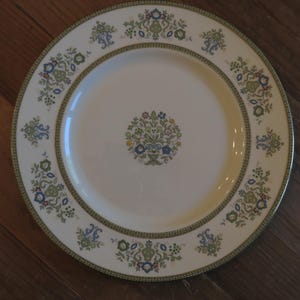 May include: A white ceramic dinner plate with a floral design. The plate features a central floral motif and a border with repeating floral patterns in green, blue, and yellow. The plate is set on a wooden surface.