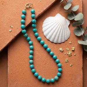 May include: A turquoise beaded necklace with a gold clasp. The necklace is displayed with a white seashell, eucalyptus sprigs, and gold leaf flakes on a terracotta-colored surface. Jewelry.