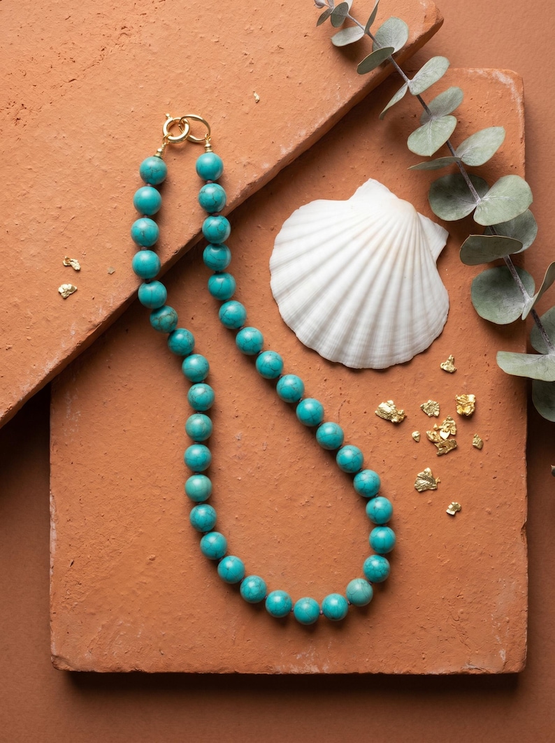May include: A turquoise beaded necklace with a gold clasp. The necklace is displayed with a white seashell, eucalyptus sprigs, and gold leaf flakes on a terracotta-colored surface. Jewelry.