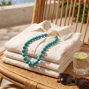 May include: A stack of folded white linen shirts with a turquoise beaded necklace and gold clasp. A glass of water with a lime wedge and tortoiseshell sunglasses rest on a woven chair. The scene is set outdoors with a view of the ocean.