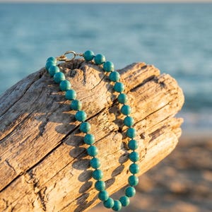 May include: A turquoise beaded necklace with a gold clasp rests on a piece of driftwood. The necklace features round, polished beads in a vibrant teal color. The background shows the ocean and sky.