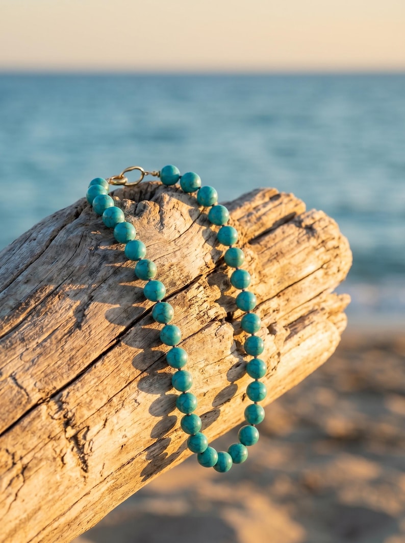 May include: A turquoise beaded necklace with a gold clasp rests on a piece of driftwood. The necklace features round, polished beads in a vibrant teal color. The background shows the ocean and sky.