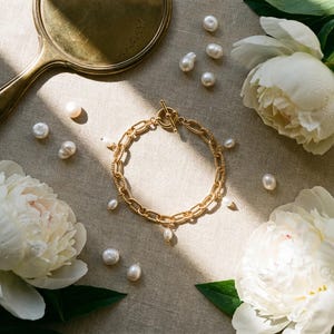 May include: A gold chain bracelet with pearl drops, alongside scattered pearls, a vintage hand mirror, and white peony flowers on a neutral linen surface. The bracelet has a toggle clasp.