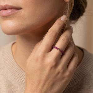 Könnte beinhalten: Nahaufnahme einer Hand, die einen Perlenring trägt. Der Ring hat kleine, runde, tiefrosa Perlen und ein paar goldfarbene Perlen. Der Ring befindet sich am Ringfinger. Die Person trägt einen beigefarbenen Pullover.