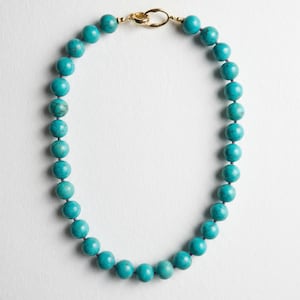 May include: A turquoise beaded necklace with a gold-colored clasp. The necklace features round, polished beads in a vibrant teal color. The clasp is a simple, elegant design, connecting the two ends of the necklace.