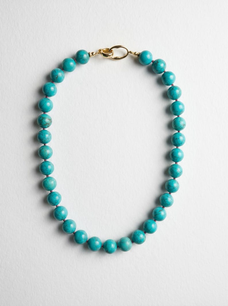 May include: A turquoise beaded necklace with a gold-colored clasp. The necklace features round, polished beads in a vibrant teal color. The clasp is a simple, elegant design, connecting the two ends of the necklace.