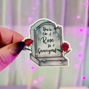 May include: A sticker featuring a tombstone design with the text "You're Like A Rose in A Graveyard." Two red roses are on either side of the tombstone. The sticker has a holographic effect.