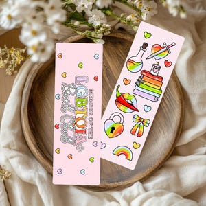 May include: Two pink bookmarks with colourful illustrations and text. One bookmark reads "LGBTQIA+ Book Club" with hearts. The other features a rainbow, books, a heart with a sword, and other symbols. Both are on a wooden tray.
