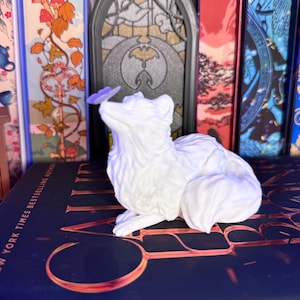 Może przedstawiać: Biała figurka lisa z motylem na nosie. Lis siedzi ze zwiniętym ogonem. Figurka umieszczona jest na książce ze złotymi literami. Tło obejmuje kolorowe grzbiety książek.