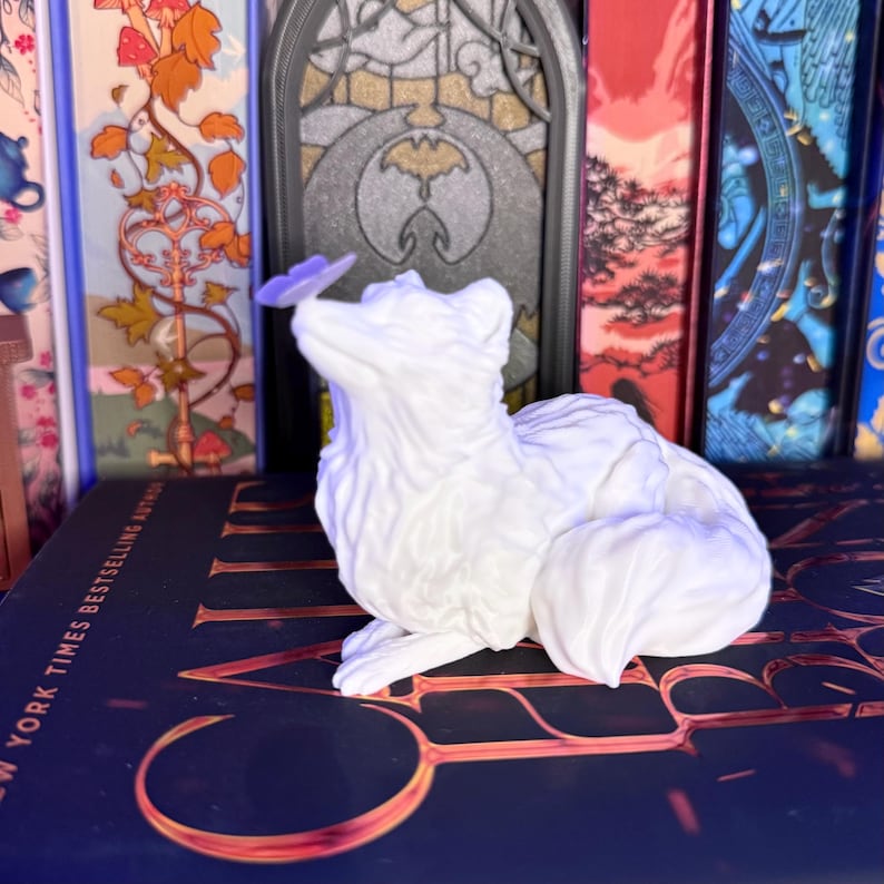 Może przedstawiać: Biała figurka lisa z motylem na nosie. Lis siedzi ze zwiniętym ogonem. Figurka umieszczona jest na książce ze złotymi literami. Tło obejmuje kolorowe grzbiety książek.