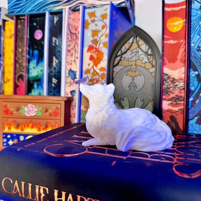 Może przedstawiać: Biała figurka lisa z motylem na nosie, spoczywająca na książce. Książka ma tytuł "Callie Hart" na okładce. W tle dekoracyjne witraże i kolekcja kolorowych książek.