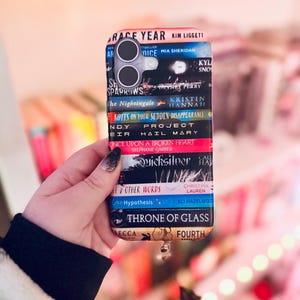 Op de afbeelding: Een telefoonhoesje met een stapel boekruggen met titels als "The Nightingale" en "Throne of Glass". De hoes heeft een zwarte rand en een camera-uitsparing. Het ontwerp bevat verschillende kleuren, waaronder rood, blauw en goud.