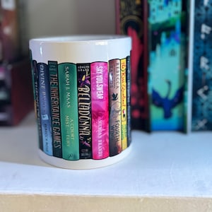 May include: White ceramic mug featuring a design of colorful book spines. The titles include "The Inheritance Games," "Belladonna," and "Say You Swear." The mug is surrounded by other books on a shelf.