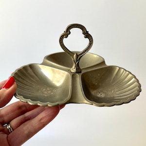 May include: A vintage, three-sectioned serving dish with a handle. The dish is made of a tarnished, silver-colored metal. Each section has a scalloped edge, and the handle has a decorative, ornate design.