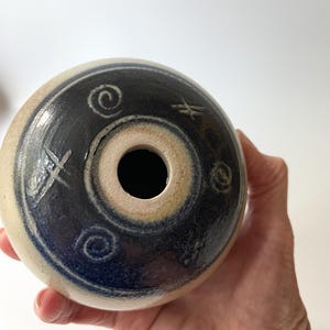 Pode incluir: Um objeto cerâmico em forma de donut com um buraco central. A superfície superior é azul escuro com desenhos brancos em espiral e em forma de cruz. A borda externa é de cor bege clara. O objeto é segurado em uma mão.