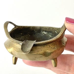 May include: A small, round, brass ashtray featuring a weathered, metallic gold finish. The vessel is supported by three short, stubby legs and includes two integrated handles on opposite sides, along with a single notched rest for a cigarette.