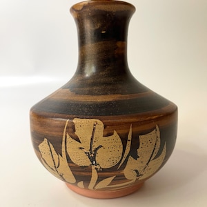 Może przedstawiać: Brązowy ceramiczny wazon z szeroką podstawą i wąską szyjką. Wazon posiada kwiatowy wzór w jaśniejszym brązowym kolorze pośrodku. Podstawa jest w kolorze jasnopomarańczowym.