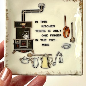 May include: Square ceramic kitchen plate with a cream background and gold trim. The plate features a black and white illustration of a stove, kettle, and kitchen utensils. Text on the plate reads: "IN THIS KITCHEN THERE IS ONLY ONE FINGER IN THE POT... MINE."