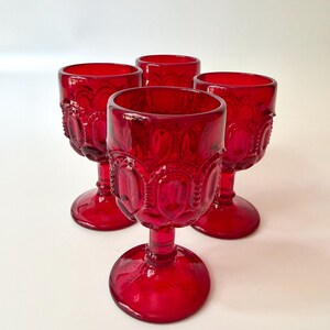 May include: Four ruby red goblets with ornate, embossed detailing. The stemware features a round base, a slender stem, and a wide bowl. The goblets are arranged on a white surface, showcasing their rich color and vintage design.