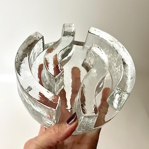 May include: Clear glass bowl with a textured surface and a unique, curved design. The bowl has a circular shape with multiple arching sections, creating an open, airy aesthetic. The glass appears thick and has a slightly irregular texture, adding visual interest.