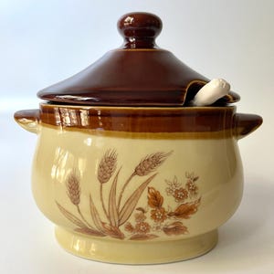 以下が含まれることがあります： 蓋付きのヴィンテージセラミック砂糖入れ。ボウルは薄い黄色で、茶色の小麦と花の模様があります。蓋とハンドルは濃い茶色です。小さな白いスプーンが縁に置かれています。