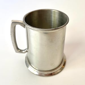 May include: A silver-colored, cylindrical tankard with a handle. The mug has a wide, circular base and a slightly flared rim. The handle is rectangular and attached to the side of the mug. The tankard is made of a shiny metal.