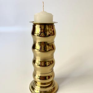 Könnte beinhalten: Ein goldfarbener Kerzenhalter aus Metall mit zylindrischem, gestapeltem Design. Oben auf dem Halter sitzt eine weiße Kerze. Der Kerzenhalter hat eine glänzende, reflektierende Oberfläche und einen runden Sockel. Die Kerze hat einen einzelnen Docht.