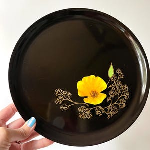May include: A round, black serving tray with a yellow poppy flower and gold-colored branch design. The tray is held by a person with blue nail polish.