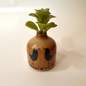 May include: A small, round, speckled tan ceramic vase featuring two black bird silhouettes painted on the front. The vase contains a small, green, artificial succulent plant with layered, pointed leaves extending from the narrow opening.