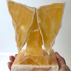 May include: An abstract sculpture crafted from golden-yellow stone. The piece features a square base, a central pillar, and wing-like forms. The stone's natural veining and white edges add to its unique aesthetic. The sculpture is a decorative art piece.