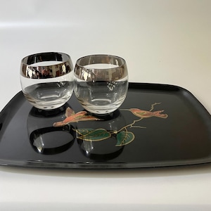 May include: A black rectangular serving tray with two clear glasses with silver rims. The tray is decorated with a bird and leaf design in shades of red, green, and gold. The glasses sit on the tray.