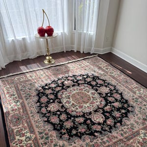 May include: A large, rectangular area rug with a central black medallion design featuring floral patterns in pink, cream, and green. The rug has a cream-colored border with floral motifs and a fringed edge. The rug is placed on a dark wood floor.