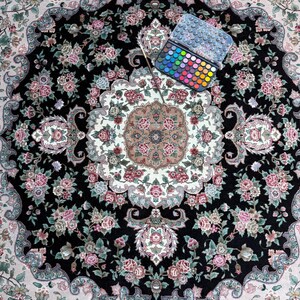 May include: An overhead shot of a detailed, round rug with a black background and floral patterns in pink, white, and green. A watercolor paint set with a brush is placed on the rug, showcasing a variety of colors.