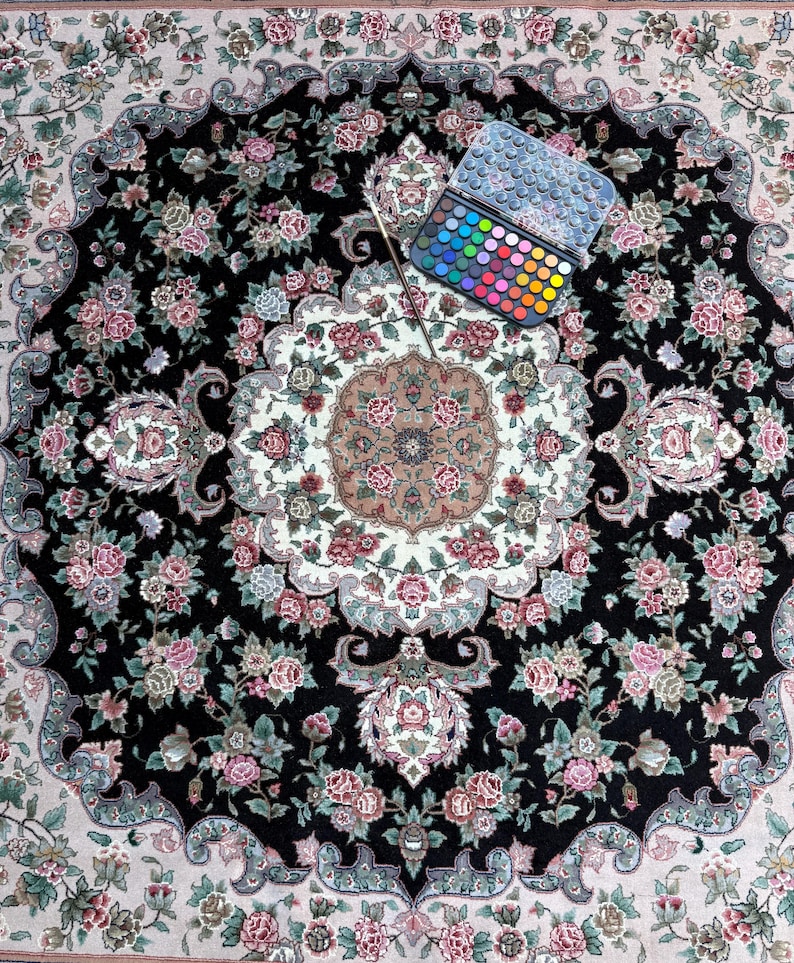May include: An overhead shot of a detailed, round rug with a black background and floral patterns in pink, white, and green. A watercolor paint set with a brush is placed on the rug, showcasing a variety of colors.