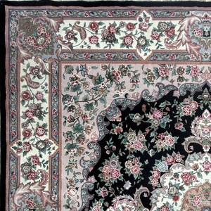 May include: Close-up of an ornate area rug with a black center and floral patterns in pink, green, and cream. The rug has a detailed border with a fringe edge, showcasing a traditional design.