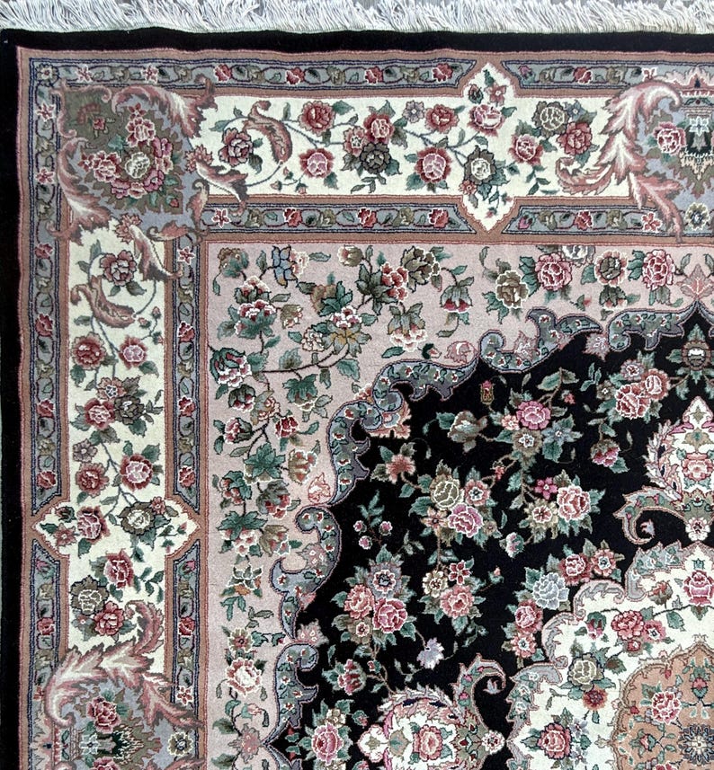 May include: Close-up of an ornate area rug with a black center and floral patterns in pink, green, and cream. The rug has a detailed border with a fringe edge, showcasing a traditional design.