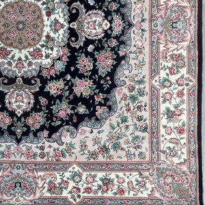 May include: Close-up of a detailed area rug with a floral pattern. The rug features a black central field with pink and green floral designs. A cream-colored border with more floral motifs and a black fringe complete the design.
