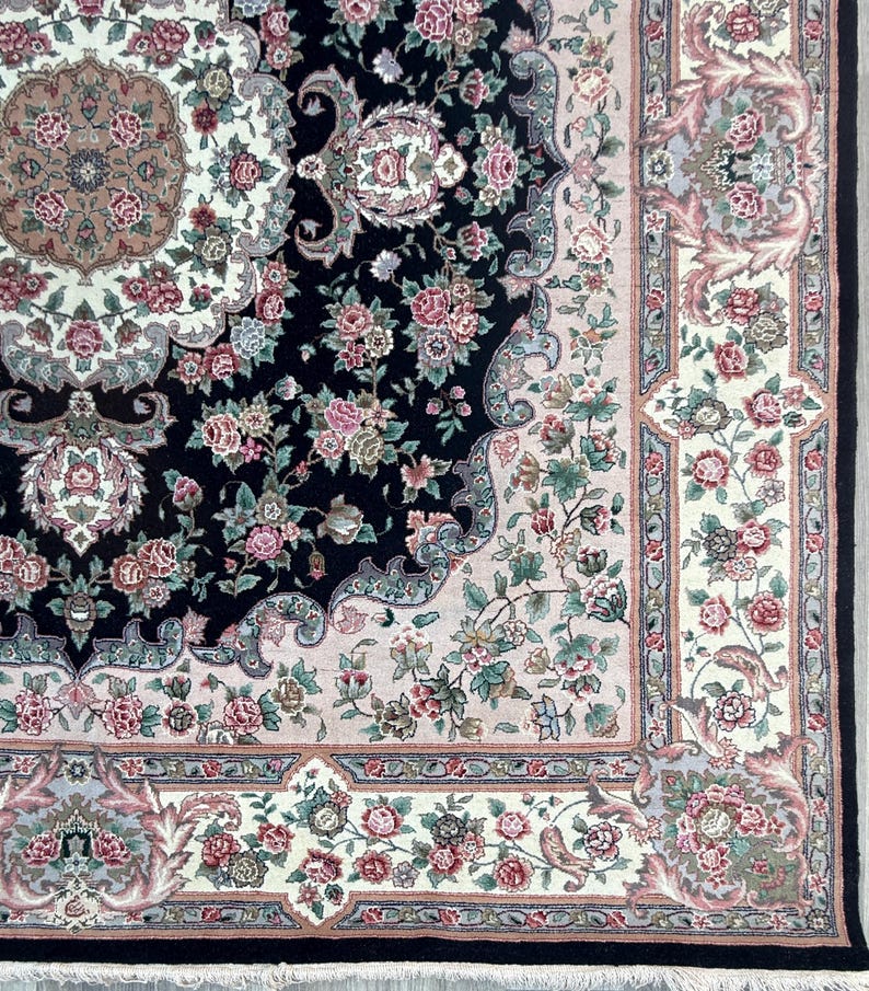 May include: Close-up of a detailed area rug with a floral pattern. The rug features a black central field with pink and green floral designs. A cream-colored border with more floral motifs and a black fringe complete the design.