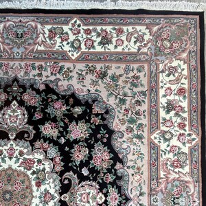 May include: Close-up of a decorative rug with a black center and floral patterns. The rug features a cream-colored border with pink and green floral designs. The edges are framed with a black border and fringe.